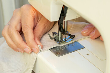 A woman sews on a sewing machine. Women's hands substitute the fabric for sewing under the foot of the sewing machine. - obrazy, fototapety, plakaty