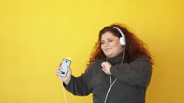 Music app. Sound energy. Fun playlist. Carefree leisure. Inspired happy chubby woman enjoying dancing in headphones listening to mp3 audio on phone isolated on yellow.