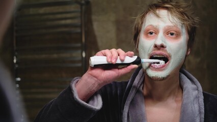 A man in a bathrobe brushes his teeth while looking in the mirror in the bathroom. The man applied...