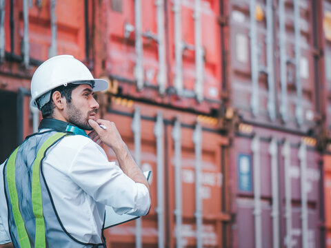 Engineer or worker wears white hardhat and reflecting cloth in front of containers cargo, carry paper files and hand touching chin with thoughtful about his job. Concept Engineering and logistic. 