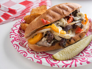 A sirloin and cheese hoagie with sauteed peppers and onions, with pickle and chips, on a paper...