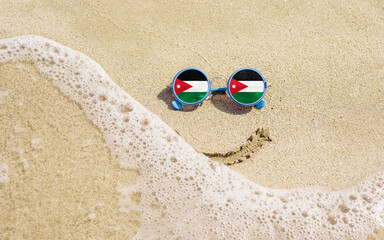 Sunglasses with flag of Jordan on a sandy beach. Nearby is a sea lightning and a painted smile. The...