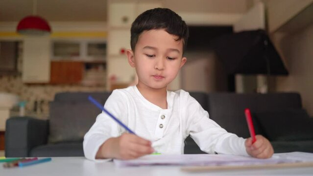 Little asian boy in white drawing using both of hands with blue and red pen on paper sitting at home