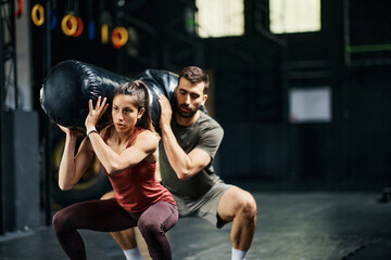 Athletic couple doing squats with sandbag on shoulders during cross training in gym.
