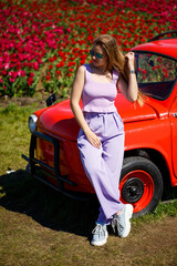 A girl model in casual clothes sits on a red car among tulip fields.