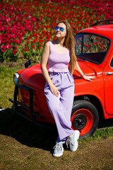 A girl model in casual clothes sits on a red car among tulip fields.