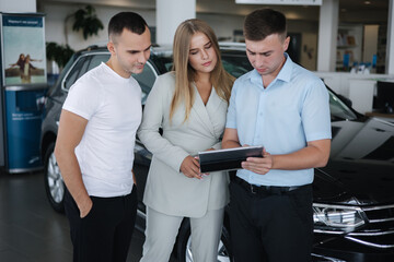 Salesperson talking with couple and help choosing new car. Man and woman in car showroom. Man using tablet to shop opportunity of different models