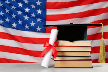 Graduation hat, books and diploma on light grey table against flag of United States. Space for text