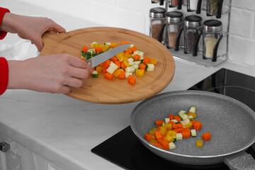 Woman putting cut vegetables onto frying pan in kitchen, closeup