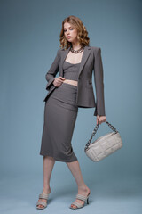 High fashion photo of a beautiful elegant young woman in pretty gray suit, jacket blazer, top, long skirt, handbag, massive chain around the neck posing on blue background. Slim figure. Monochrome