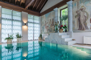 A swimming pool in a country house with large windows and frescoes on the walls.