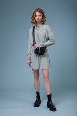 Fototapeta premium High fashion photo of a beautiful elegant young woman in pretty soft blue gray dress, boots, black handbag posing on blue background. Slim figure. Studio shot. Monochrome