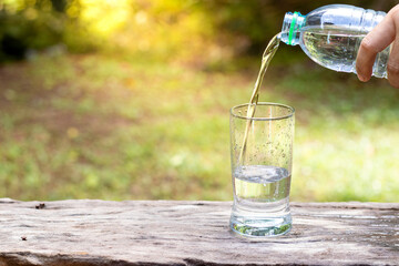 Hand holding drinking water bottle pouring water into glass on wooden table on blurred green nature background.