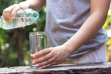 Hand holding drinking water bottle pouring water into glass on wooden table on blurred green nature background.