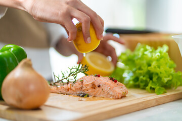 Close up of Asian woman hand squeeze lemon juice on piece of salmon steak in the kitchen for dinner at home. Happy female enjoy cooking and eating healthy food on holiday vacation