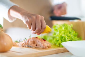 Close up of Asian woman hand squeeze lemon juice on piece of salmon steak in the kitchen for dinner at home. Happy female enjoy cooking and eating healthy food on holiday vacation
