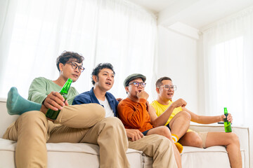 Group of Asian man friends sitting on sofa watching soccer game competition on television with drinking beer together at home. Happy male soccer fans cheering victory football team victory sport match