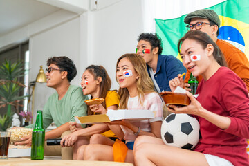 Group of Asian people friends sitting on sofa watching soccer games competition on television with eating food together at home. Man and woman sport fans celebrating sport team victory national match