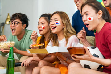 Group of Asian people friends sitting on sofa watching soccer games competition on television with eating food together at home. Man and woman sport fans celebrating sport team victory national match