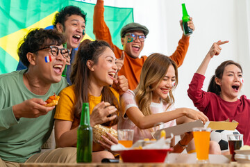 Group of Asian people friends sitting on sofa watching soccer games competition on television with eating food together at home. Man and woman sport fans celebrating sport team victory national match