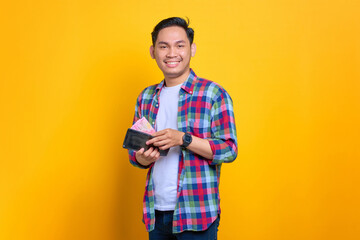 Cheerful young Asian man in plaid shirt showing wallet with cash money isolated on yellow background