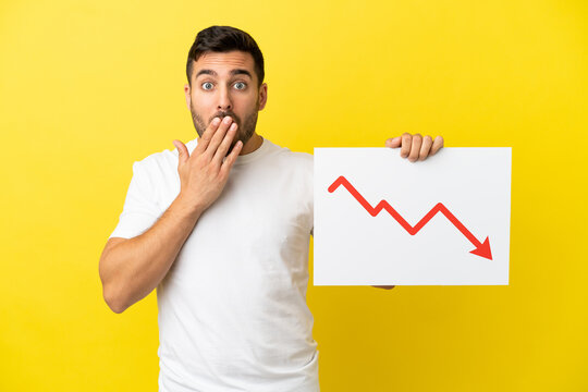 Young handsome caucasian man isolated on yellow background holding a sign with a decreasing statistics arrow symbol with surprised expression
