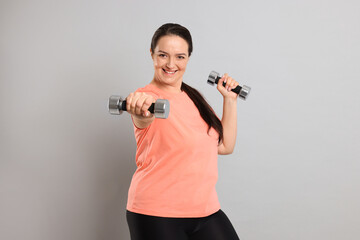 Happy overweight woman doing exercise with dumbbells on grey background
