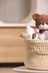 Knitted basket with baby cosmetic products, bath accessories and toy bear on white table indoors - obrazy, fototapety, plakaty