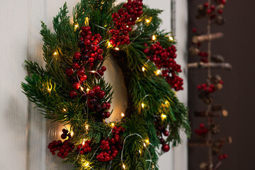 Beautiful Christmas wreath with red berries and fairy lights hanging on white door, closeup. Space...