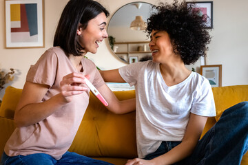 Smiling young woman shows girlfriend positive pregnancy test. Happy multiracial lesbian couple find...