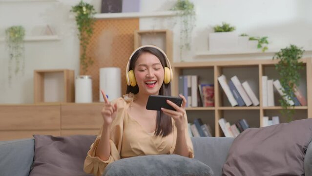 Asian young woman in yellow headphones sitting in couch in living room at home listening favorite Music and dancing on sofa, move arms. Relax and enjoying lazy weekend hobby time alone at home.