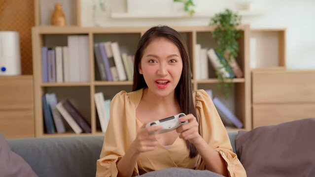 Young asian woman playing game online at home. Gamer female controlling joystick for Video game console or computer game in front of camera. Cheerful girl celebrating gaming victory and success.