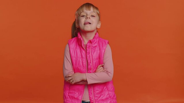 Cheerful funny girl in pink vest showing tongue making faces at camera, fooling around, joking, aping with silly face, teasing. Young little blonde child kid isolated alone on orange studio background