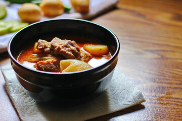Massaman curry in black crockery served with boiled potatoes and chili on a brown wooden table.