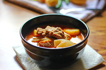 Massaman curry in black crockery served with boiled potatoes and chili on a brown wooden table.