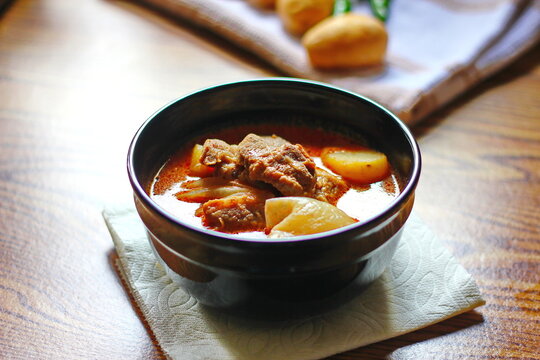 Massaman curry in black crockery served with boiled potatoes and chili on a brown wooden table.