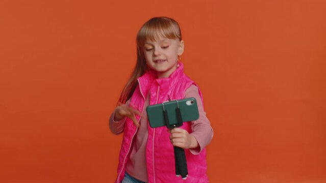 Girl child toddler blogger taking selfie on mobile phone selfie stick, communicating video call online with subscribers. Young little children kid teen isolated alone on orange studio wall background