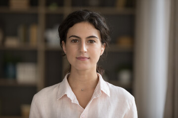 Head shot serious good-looking young 20s woman in casual elegant shirt standing alone in cozy living room staring at camera feels confident pose indoor. Housemaid or real-estate agent portrait concept