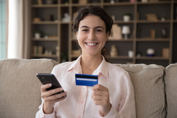 Portrait of e-commerce services client concept. 25s woman sit on sofa smile look at camera holds smartphone and credit card, make order, buy goods and e-services on internet, pay bills via e-bank app