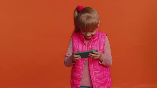 Worried funny addicted girl in vest enthusiastically playing racing video games on mobile phone. Young little child kid using smartphone gadget app drive simulator isolated on orange studio background