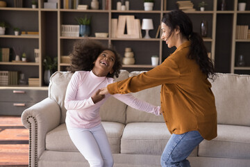 Cute little girl and mom hold hands dancing in living room. Young woman preschool daughter moving to favourite music looking carefree, feel overjoyed. Family enjoy hobby, spend leisure at home concept