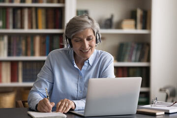 Happy elder student in headphones watching learning webinar on laptop, virtual workshop, writing notes. Getting knowledge, new profession, studying online from home, taking e-learning course