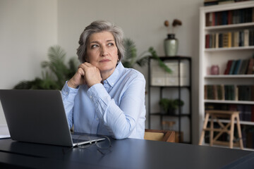 Thoughtful serious mature grey haired business lady sitting at work office table with laptop,...