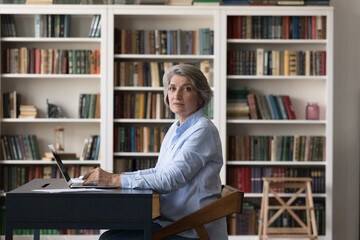 Serious senior university professor woman, mature college student working at laptop in library, looking at camera from workplace with bookshelves in background. Indoor portrait