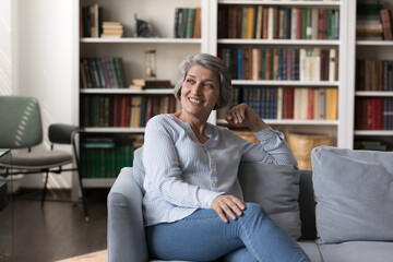 Obraz premium Happy cheerful senior 60s woman sitting on comfortable sofa in living room, looking away, smiling at good thoughts, thinking over future retirement plans, insurance terms, enjoying home leisure time