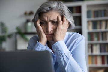 Frustrated senior retired woman touching head, looking at laptop screen with despair, getting bad...
