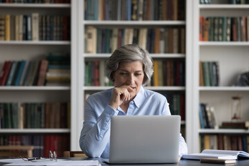 Positive focused senior business lady using laptop at table in home office, library with...