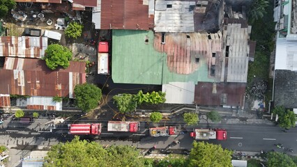Aerial view of firefighters extinguishing the ruins of a burning furniture factory building with a collapsed roof and billowing smoke.