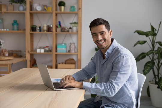 Happy confident businessman, entrepreneur, small business owner portrait at home office workplace. Young handsome freelance employee, student using laptop computer, looking at camera, smiling
