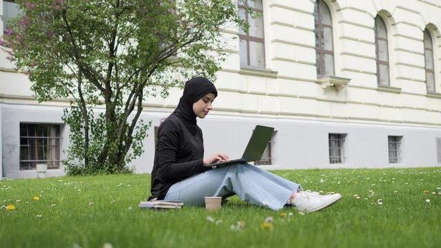 Beautiful muslim arab islamic student girl in hijab studies outside. Woman freelancer user uses laptop sitting on green grass in park near university working online studying remotely chatting outdoor.